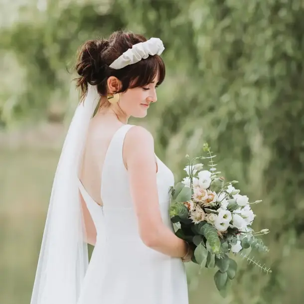 Moderne Braut mit Haarreife Schleier und Brautstrauß schaut in Richtung des Straußes vor grüner Kulisse an der Donau