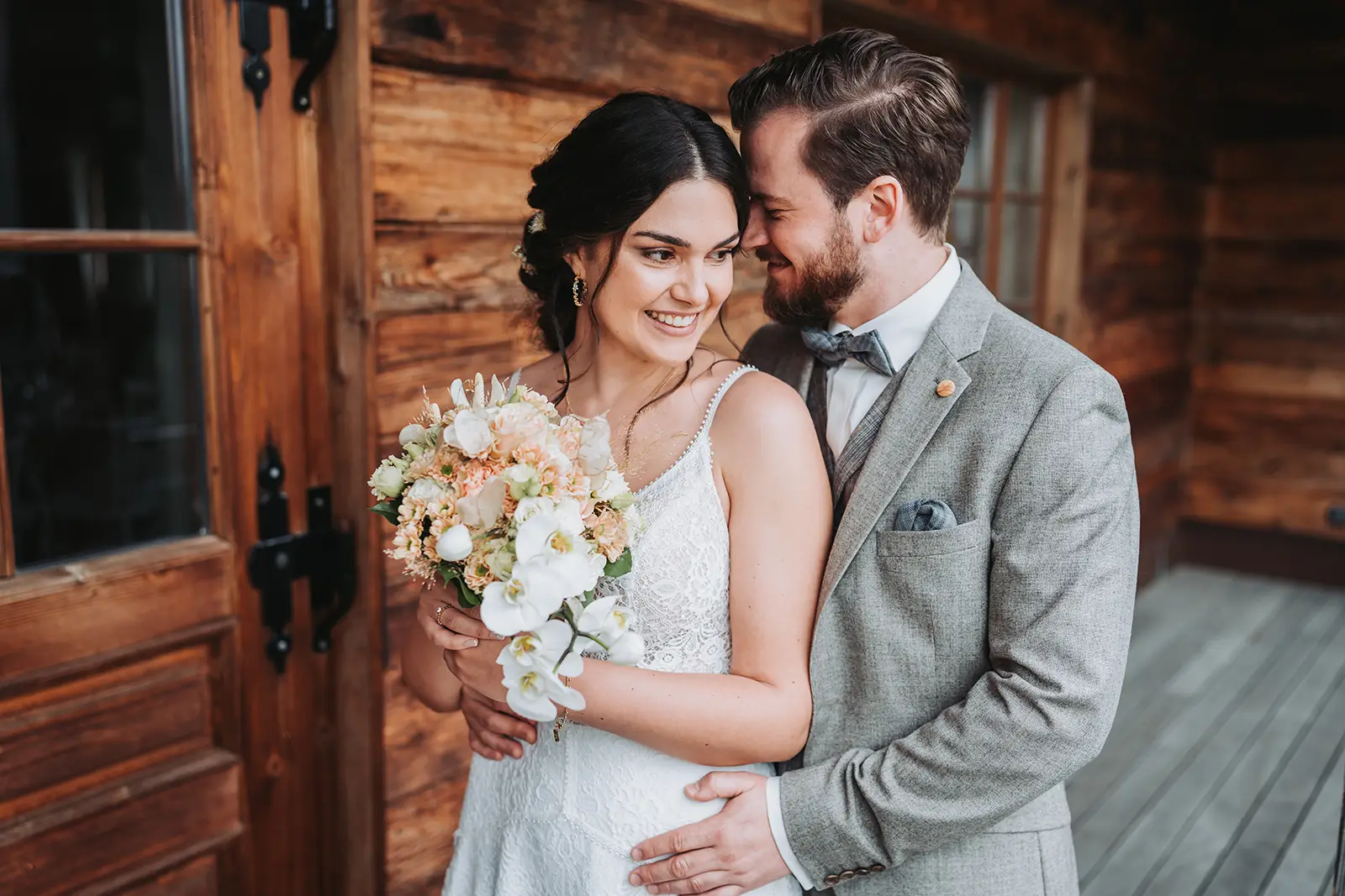 Bräutigam in grauem Anzug umarmt seine Braut mit Blumenstrauß leicht seitlich aufgenommen vor der rustikalen Werkstatt der Scheunenwirtin in Bartholomä